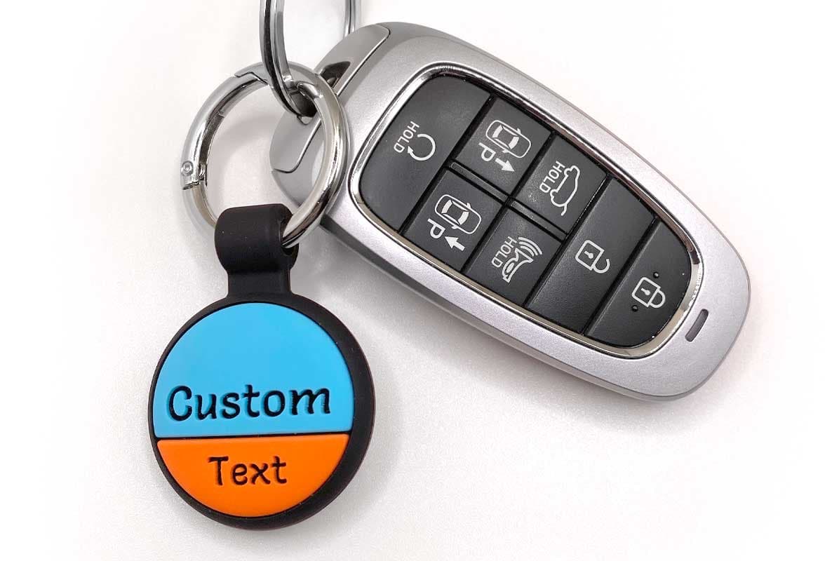 Front view of blue-orange AirTag keychain with two-line engraved Custom Text, metal split ring and attached car key on white