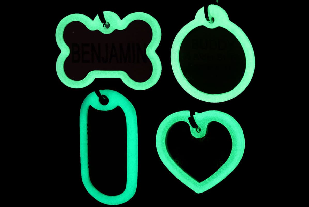 Front view of four glow-in-the-dark pet tag silencers (bone, round, rectangle, heart) on black background