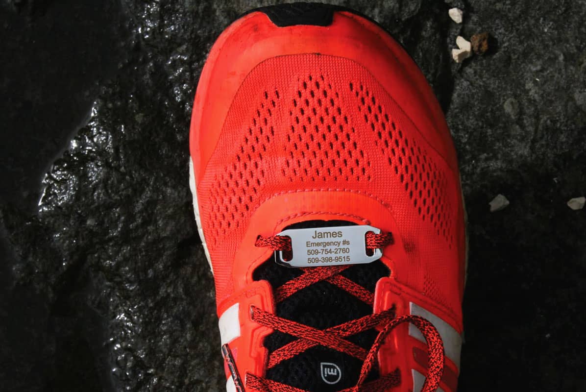 Top view of Shoe ID Tag on red running shoe laces showing engraved name James and two emergency phone numbers